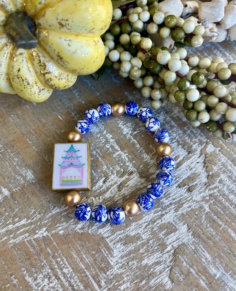 K&ouml;nnte beinhalten: Ein Armband mit blauen und wei&szlig;en Porzellanperlen und goldfarbenen Perlen. Es hat einen rechteckigen Anh&auml;nger mit einem Pagoden-Design in Rosa, Gr&uuml;n und Blau. Das Armband wird auf einer Holzoberfl&auml;che pr&auml;sentiert.
