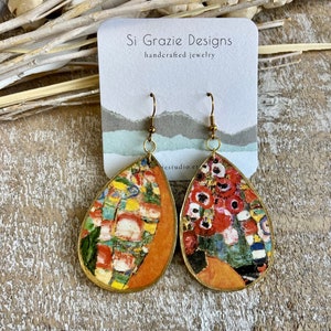 May include: Teardrop-shaped earrings with a colorful abstract design, featuring gold-tone hooks. The earrings have a gold border and a vibrant mix of colors, including red, yellow, and blue. The earrings are displayed on a white card with the text "Si Grazie Designs handcrafted jewelry."