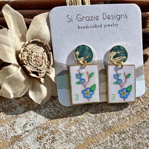May include: Handcrafted jewelry earrings featuring a floral design. The earrings have a gold border and a white background with a blue floral design and the number 3. The earrings are attached to a gold-colored circular top.