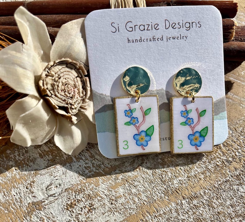 May include: Handcrafted jewelry earrings featuring a floral design. The earrings have a gold border and a white background with a blue floral design and the number 3. The earrings are attached to a gold-colored circular top.
