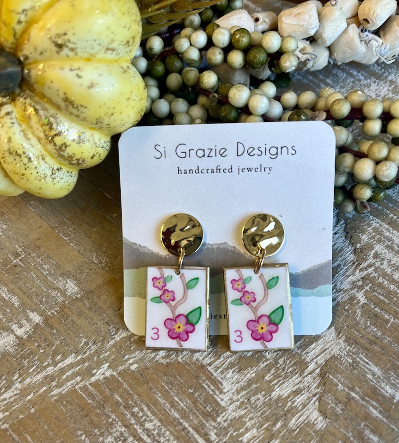 May include: Handcrafted jewelry earrings with gold-tone hammered disc tops and rectangular white pendants. The pendants feature pink flowers, green leaves, and the number 3. The earrings are from Si Grazie Designs.