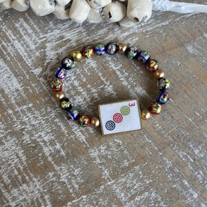 Op de afbeelding: Een armband met kleurrijke glazen kralen en goudkleurige kralen. De armband bevat een vierkante bedel met het nummer "3" en drie cirkelvormige symbolen. De armband wordt gepresenteerd op een houten oppervlak.