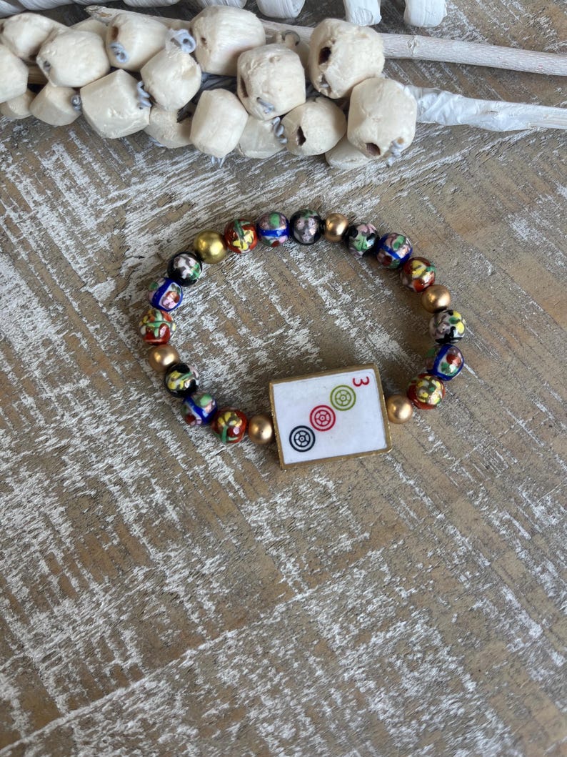 Op de afbeelding: Een armband met kleurrijke glazen kralen en goudkleurige kralen. De armband bevat een vierkante bedel met het nummer "3" en drie cirkelvormige symbolen. De armband wordt gepresenteerd op een houten oppervlak.