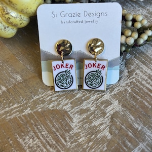 May include: Gold-toned stud earrings with rectangular white pendants featuring the word "JOKER" and a green design. The earrings are handcrafted jewelry, as indicated by the tag in the background.