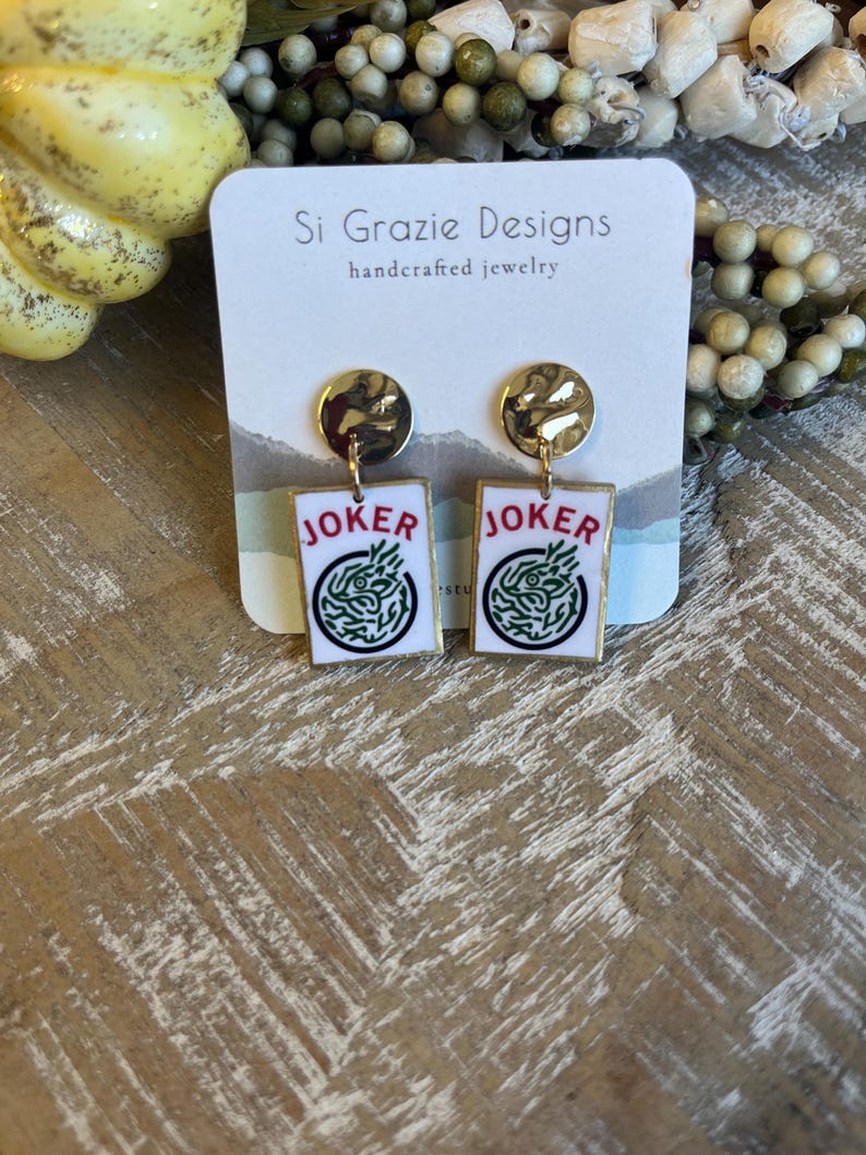 May include: Gold-toned stud earrings with rectangular white pendants featuring the word "JOKER" and a green design. The earrings are handcrafted jewelry, as indicated by the tag in the background.