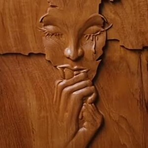 Può includere: Una scultura in legno del viso di una donna con gli occhi chiusi e una singola lacrima che le scorre sulla guancia. La scultura è scolpita in uno stile realistico e l'espressione della donna è di tristezza.