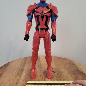 May include: Red and blue action figure of Spider-Man wearing a red and black armored suit. The figure is standing on a wooden surface with a ruler for scale.