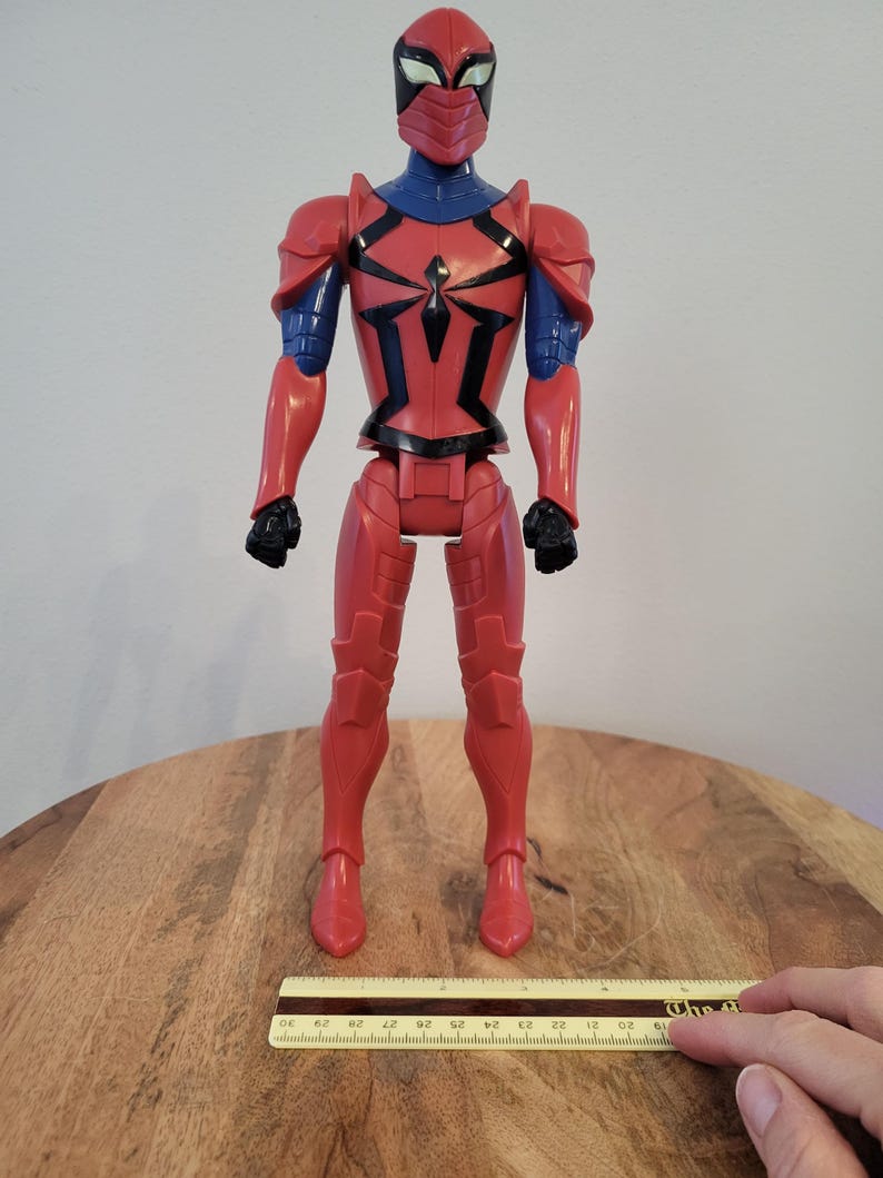 May include: Red and blue action figure of Spider-Man wearing a red and black armored suit. The figure is standing on a wooden surface with a ruler for scale.