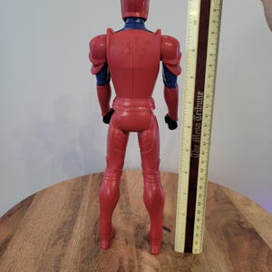 May include: Red and blue action figure wearing armor, standing on a wooden surface. The figure is approximately 12 inches tall.