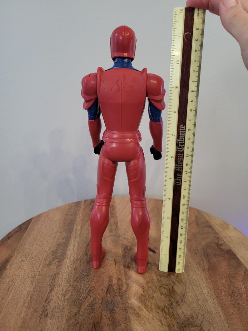 May include: Red and blue action figure wearing armor, standing on a wooden surface. The figure is approximately 12 inches tall.