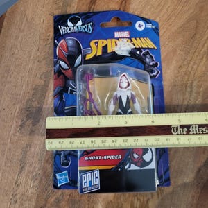 K&ouml;nnte beinhalten: Eine Ghost-Spider-Actionfigur in einer Venom Versus Marvel Spider-Man-Verpackung. Die Figur ist wei&szlig;, schwarz und lila. Die Verpackung ist blau mit dem Marvel Spider-Man-Logo und dem Text "EPIC WORLD OF ACTION". Die Verpackung ist ca. 15 cm lang.