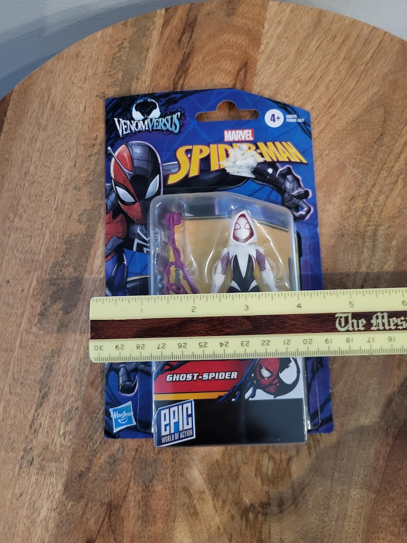 K&ouml;nnte beinhalten: Eine Ghost-Spider-Actionfigur in einer Venom Versus Marvel Spider-Man-Verpackung. Die Figur ist wei&szlig;, schwarz und lila. Die Verpackung ist blau mit dem Marvel Spider-Man-Logo und dem Text "EPIC WORLD OF ACTION". Die Verpackung ist ca. 15 cm lang.