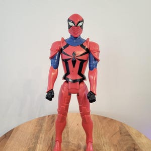 May include: Red and blue action figure of a superhero wearing a red and black suit with a spider symbol on the chest. The figure is wearing a red helmet with a blue mask.