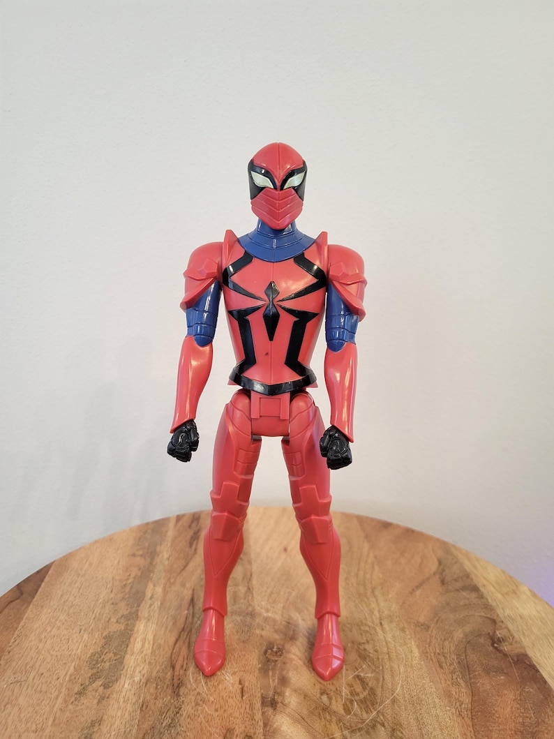 May include: Red and blue action figure of a superhero wearing a red and black suit with a spider symbol on the chest. The figure is wearing a red helmet with a blue mask.