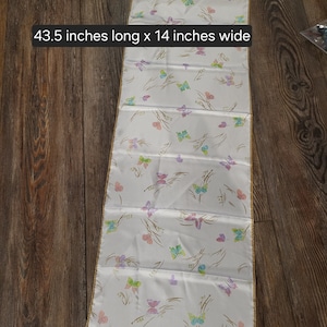 May include: White table runner with a gold trim and a butterfly pattern. The runner is 43.5 inches long and 14 inches wide.