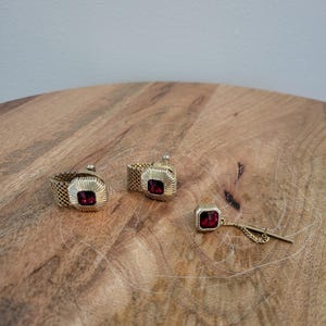 May include: A set of two gold-tone mesh cufflinks and a matching tie tack, each featuring a square-cut deep red gemstone set within a ribbed gold-tone frame. The cufflinks have a flexible mesh band, and the tie tack includes a short chain.