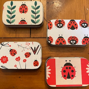 May include: Four small rectangular metal tins with different ladybug designs. The tins are white with red and black ladybugs and green leaves. The tins are on a wooden surface.