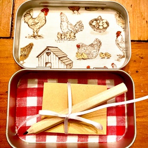 May include: A small metal tin with a red and white checkered pattern lining. The tin contains a brown paper note and a wooden pencil tied together with a white ribbon. The lid of the tin has a white background with a brown and white chicken and rooster pattern.