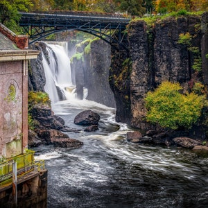 May include: A view of a waterfall cascading over rocks with a bridge arching over the top. A brick building with a green roof is on the left side of the image.