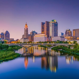 May include: A panoramic view of the Columbus, Ohio skyline at dusk. The city's skyscrapers are illuminated, reflecting in the Scioto River. A bridge spans the river in the foreground, with lush green trees lining the banks.