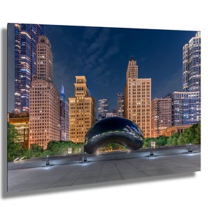 May include: A night-time cityscape featuring illuminated skyscrapers and a large, reflective, bean-shaped sculpture. The buildings are lit with warm tones against a dark blue sky, with the sculpture in the foreground.