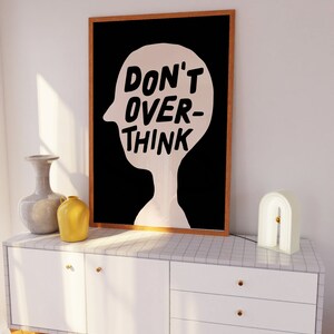 May include: A black and white framed print with the text "DON'T OVER-THINK" inside a silhouette of a head. The print is hanging on a white wall above a white dresser with three drawers.