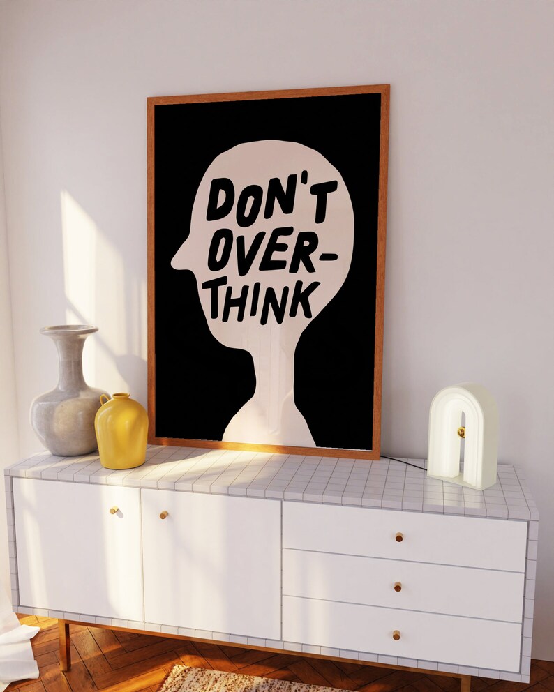 May include: A black and white framed print with the text "DON'T OVER-THINK" inside a silhouette of a head. The print is hanging on a white wall above a white dresser with three drawers.