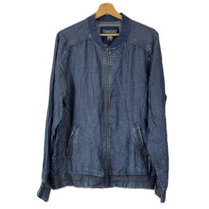 May include: A blue denim bomber jacket with a front zip fastening, ribbed collar, cuffs, and hem. The jacket features two side pockets and a label at the inner neckline with the text "TRANQUILLO" and a small green elephant graphic icon.