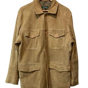 Puede incluir: Chaqueta de ante color canela con cuatro bolsillos delanteros y botones. La chaqueta tiene cuello y está colgada de una percha de madera. La marca "SPRINGFIELD" es visible en una etiqueta dentro del cuello.