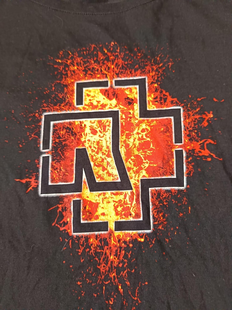 May include: Black t-shirt featuring a white and black graphic of a cross-like symbol with the letters "R" and "W" inside, set against a fiery orange and yellow background. The design has a distressed, grunge aesthetic.