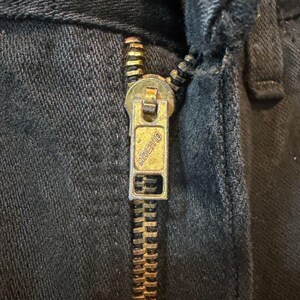 K&ouml;nnte beinhalten: Nahaufnahme eines schwarzen Denim-Kleidungsst&uuml;cks mit einem Messingrei&szlig;verschluss. Der Rei&szlig;verschlussanh&auml;nger ist rechteckig und tr&auml;gt den eingepr&auml;gten Markennamen "Premo". Die Rei&szlig;verschlussz&auml;hne und der Anh&auml;nger weisen Gebrauchsspuren auf. Der Stoff ist dunkel und strukturiert.
