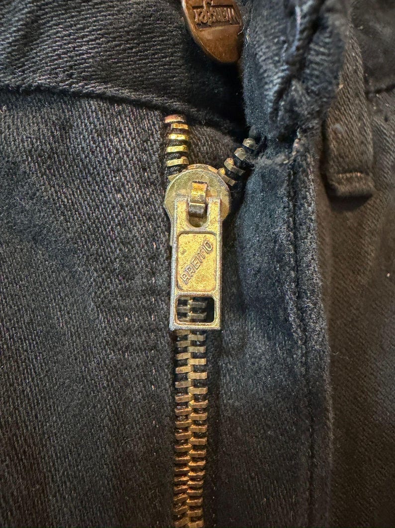 K&ouml;nnte beinhalten: Nahaufnahme eines schwarzen Denim-Kleidungsst&uuml;cks mit einem Messingrei&szlig;verschluss. Der Rei&szlig;verschlussanh&auml;nger ist rechteckig und tr&auml;gt den eingepr&auml;gten Markennamen "Premo". Die Rei&szlig;verschlussz&auml;hne und der Anh&auml;nger weisen Gebrauchsspuren auf. Der Stoff ist dunkel und strukturiert.