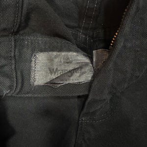 K&ouml;nnte beinhalten: Nahaufnahme einer schwarzen Jeanshose mit sichtbarem Rei&szlig;verschluss und dem Wrangler-Etikett. Das Etikett ist grau und rechteckig, mit dem Wrangler-Logo und Text. Die Hose besteht aus einem dunklen, strapazierf&auml;higen Stoff.