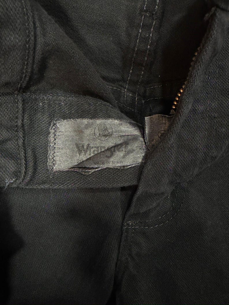 K&ouml;nnte beinhalten: Nahaufnahme einer schwarzen Jeanshose mit sichtbarem Rei&szlig;verschluss und dem Wrangler-Etikett. Das Etikett ist grau und rechteckig, mit dem Wrangler-Logo und Text. Die Hose besteht aus einem dunklen, strapazierf&auml;higen Stoff.