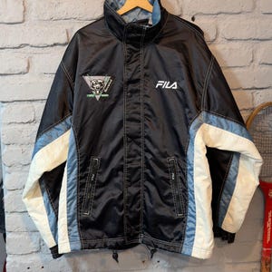 Op de afbeelding: Zwarte Fila jas met witte en lichtblauwe accenten. Voorzien van een hoge kraag, drukknoopsluiting en het Fila-logo op de borst. Inclusief een patch met een berenafbeelding. Een vintage sportjas.
