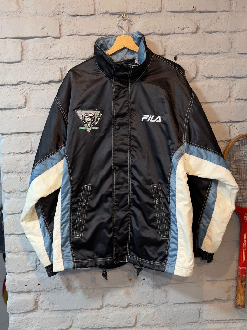 Op de afbeelding: Zwarte Fila jas met witte en lichtblauwe accenten. Voorzien van een hoge kraag, drukknoopsluiting en het Fila-logo op de borst. Inclusief een patch met een berenafbeelding. Een vintage sportjas.