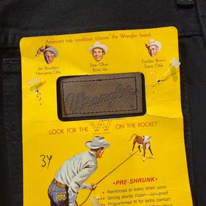 K&ouml;nnte beinhalten: Nahaufnahme eines gelben Wrangler Jeans-Etiketts auf schwarzem Denim. Das Etikett zeigt Illustrationen von Cowboys und den Text "America's top cowboys choose the Wrangler brand". Das Etikett enth&auml;lt auch den Text "PRE-SHRUNK" und "Wrangler."
