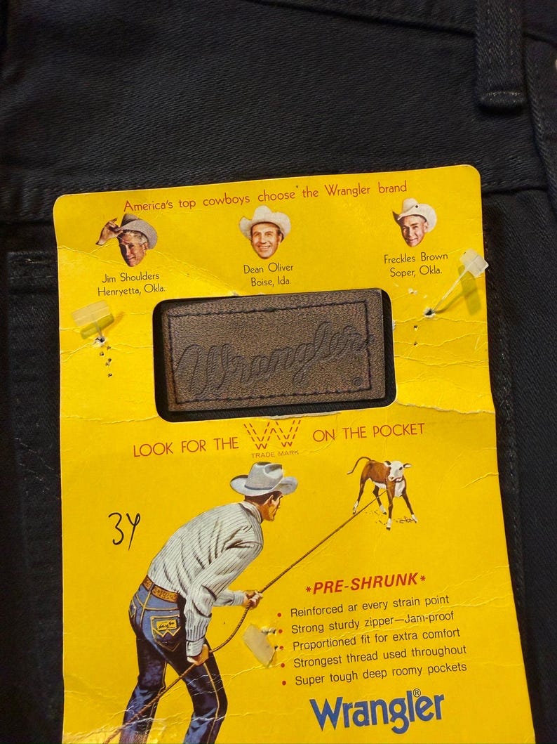 K&ouml;nnte beinhalten: Nahaufnahme eines gelben Wrangler Jeans-Etiketts auf schwarzem Denim. Das Etikett zeigt Illustrationen von Cowboys und den Text "America's top cowboys choose the Wrangler brand". Das Etikett enth&auml;lt auch den Text "PRE-SHRUNK" und "Wrangler."