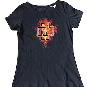 May include: Black t-shirt featuring a graphic design with a cross-like symbol in red, orange, and yellow. The design has a fiery, textured appearance. The shirt has a crew neck and short sleeves.