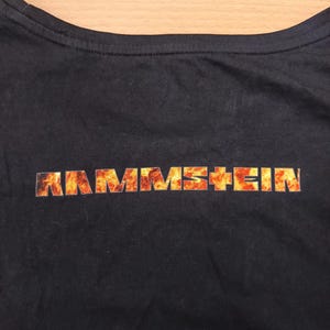 May include: Black t-shirt featuring the band name "RAMMSTEIN" in a fiery orange and yellow font across the chest. The shirt has a crew neck and is made of a dark fabric.