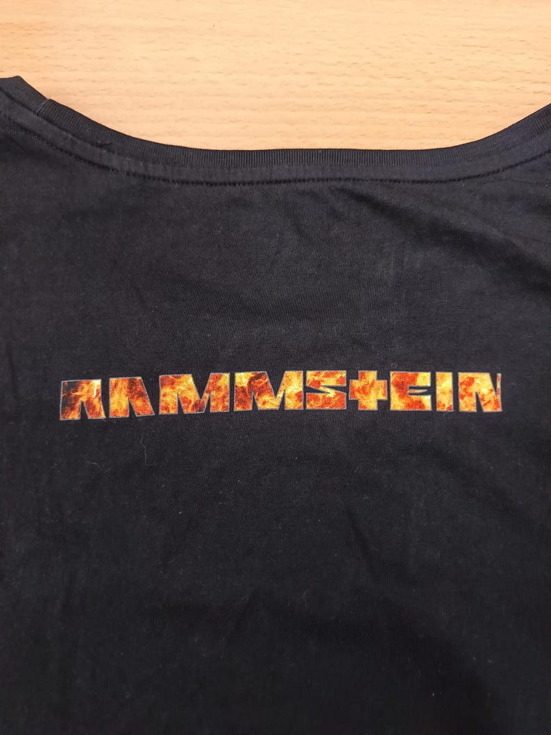 May include: Black t-shirt featuring the band name "RAMMSTEIN" in a fiery orange and yellow font across the chest. The shirt has a crew neck and is made of a dark fabric.