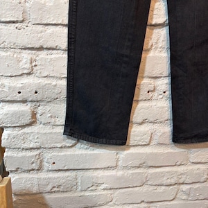 K&ouml;nnte beinhalten: Nahaufnahme einer schwarzen Jeans aus Denim vor einer wei&szlig;en Backsteinmauer. Die Jeans hat eine einfarbige schwarze Farbe, mit sichtbaren N&auml;hten und einem klassischen geraden Bein. Der Stoff scheint robust und gut verarbeitet zu sein, geeignet f&uuml;r den Alltag.