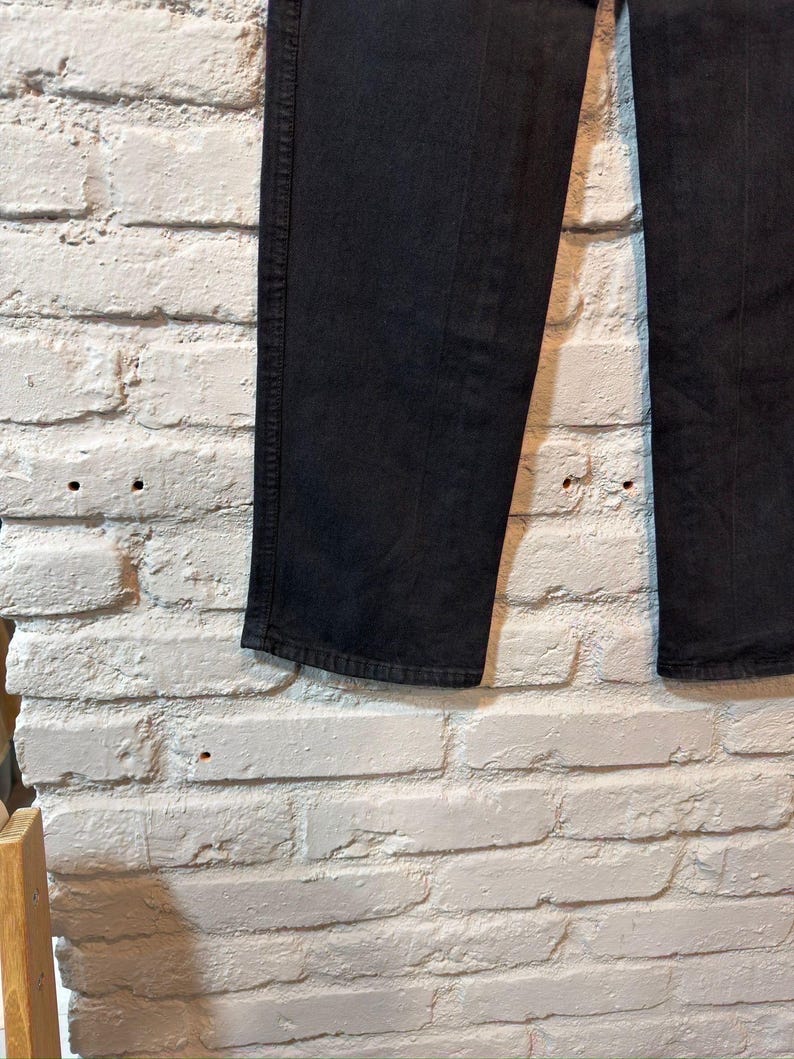 K&ouml;nnte beinhalten: Nahaufnahme einer schwarzen Jeans aus Denim vor einer wei&szlig;en Backsteinmauer. Die Jeans hat eine einfarbige schwarze Farbe, mit sichtbaren N&auml;hten und einem klassischen geraden Bein. Der Stoff scheint robust und gut verarbeitet zu sein, geeignet f&uuml;r den Alltag.