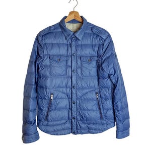Könnte beinhalten: Blaue Steppjacke mit Knopfleiste, zwei Brusttaschen und zwei Reißverschlusstaschen. Die Jacke hängt an einem Holzbügel vor weißem Hintergrund. Die Jacke hat einen Kragen und lange Ärmel.