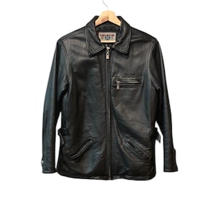 May include: Black leather jacket with a zipper closure, two front pockets, and a collared neckline. The jacket has the text "BOMB BOOGIE" on the label and a small logo on the chest. The jacket is hanging on a wooden hanger.