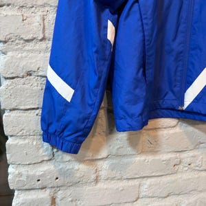 Puede incluir: Chaqueta azul brillante con rayas blancas en las mangas. La chaqueta tiene una cremallera completa y un bolsillo. El material parece ligero, adecuado para usar en capas.