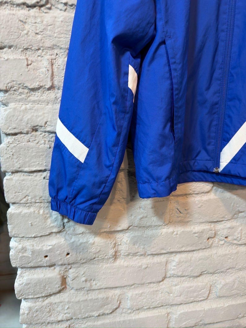 Puede incluir: Chaqueta azul brillante con rayas blancas en las mangas. La chaqueta tiene una cremallera completa y un bolsillo. El material parece ligero, adecuado para usar en capas.