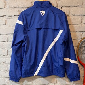 Puede incluir: Chaqueta deportiva azul con rayas blancas y un logotipo en la espalda. La chaqueta est&aacute; colgada de una percha de madera contra una pared de ladrillo. La chaqueta tiene cuello alto y cremallera.