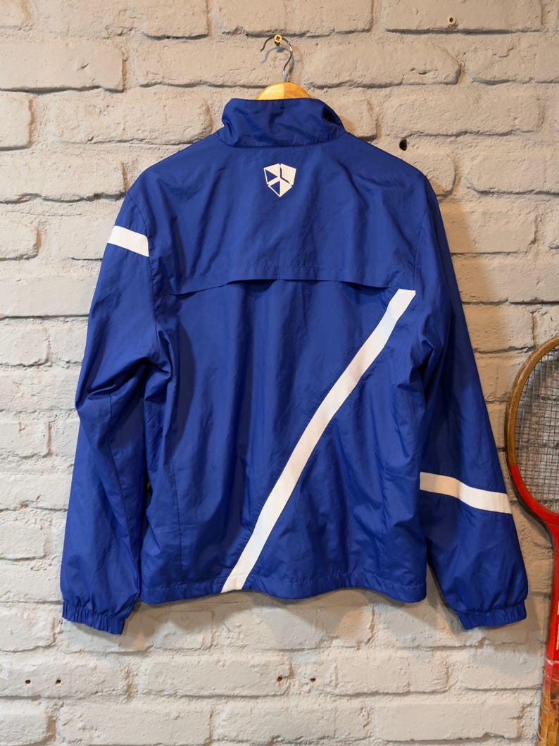 Puede incluir: Chaqueta deportiva azul con rayas blancas y un logotipo en la espalda. La chaqueta est&aacute; colgada de una percha de madera contra una pared de ladrillo. La chaqueta tiene cuello alto y cremallera.