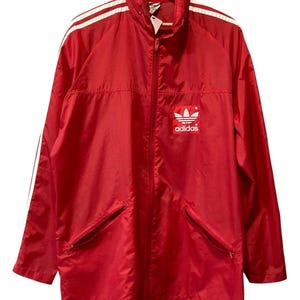 Puede incluir: Chaqueta deportiva Adidas roja con rayas blancas en las mangas y el logo de Adidas en el pecho. La chaqueta tiene cremallera completa, dos bolsillos delanteros y cuello alzado. Colgada de una percha de madera.